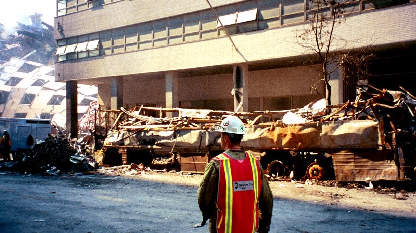 Man in helmet and orange visibility vest in front of a crumbled building